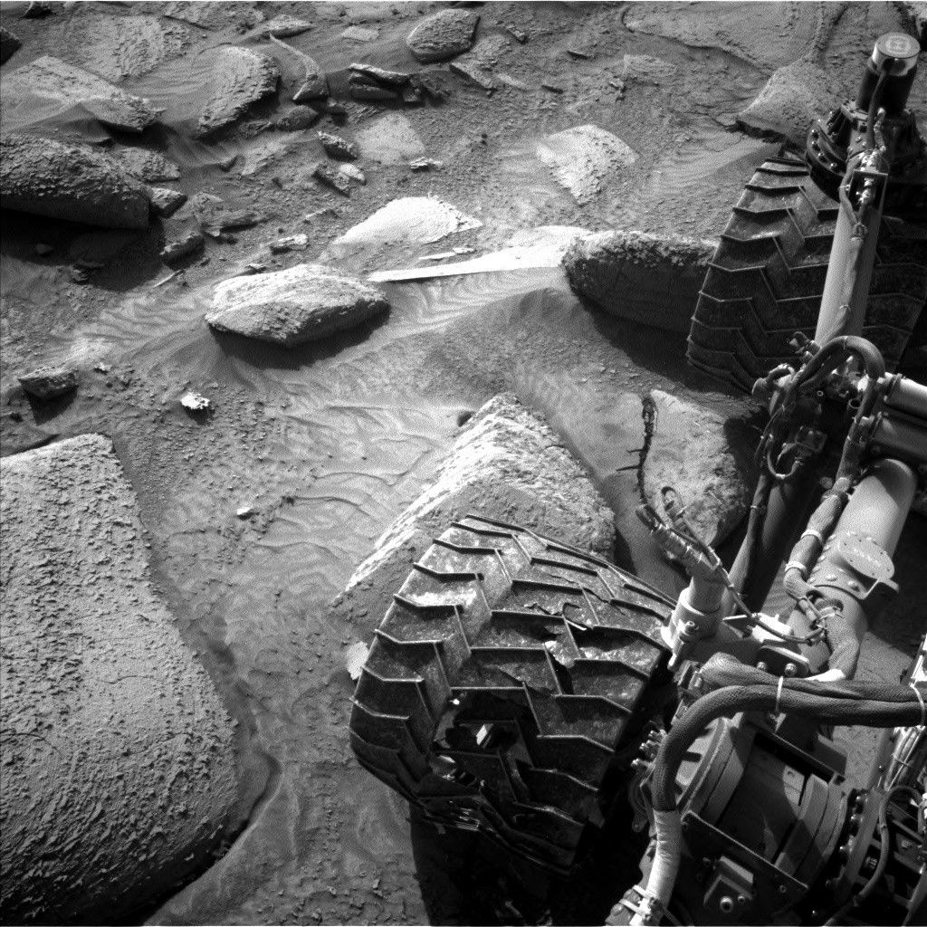 Марсоход NASA «Кьюриосити» сделал это изображение, на котором видно, как его колесо неуклюже балансирует на вершине одного из камней в этом месте, а также текстуру слоёв сульфатных пород. 28 февраля 2025 года, в 00:34:10 по всемирному координированному времени, марсоход использовал левую навигационную камеру (Navcam), одну из пары стереокамер по обе стороны от мачты марсохода, чтобы сделать это изображение.