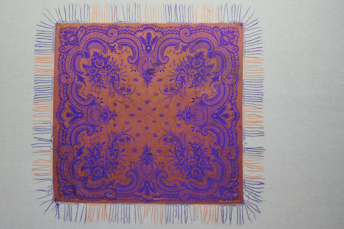 АМДЗ КП-15282. Т-2817 ШАЛЬ. Конец ХIХ - начало ХХ в. Ткань шелковая, фабричное производство. 151х144. На оранжевом фоне - тканый узор фиолетового цвета. В среднике - мелкие цветы и букетики. В широкой кайме - крупные завитки-волюты с двойными фестонами по нижнему краю и букеты из пышных цветов, трав и листьев. По краям привязаны кисти из шелковых нитей фиолетового и оранжевого цвета. Экспедиция в Лешуконский район, 2002.