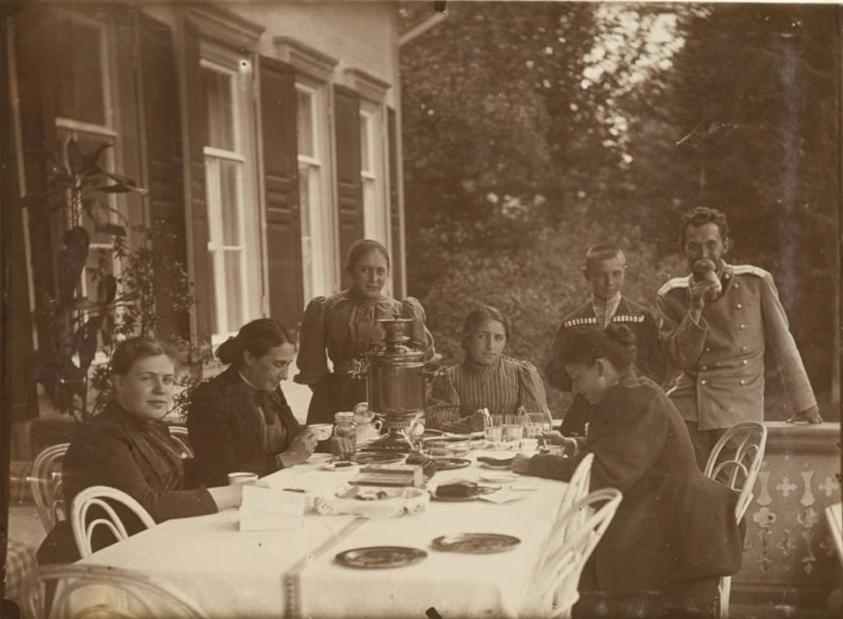 Фотография. Группа за столом на террасе абрамцевского дома. Слева направо за столом сидят: Наталья Яковлевна Давыдова - художница, одна из руководителей абрамцевской столярной мастерской, Е.Г. Мамонтова, А.С. Мамонтова, М.Ф. Мамонтова - Якунчикова. Стоят: В.С Мамонтова, Д.К. Арцыбушев (сын К.Д. Арцыбушева, сотрудника С.И. Мамонтова по его промышленным делам), С.С. Мамонтов. 1890-е // Музей-заповедник "Абрамцево"