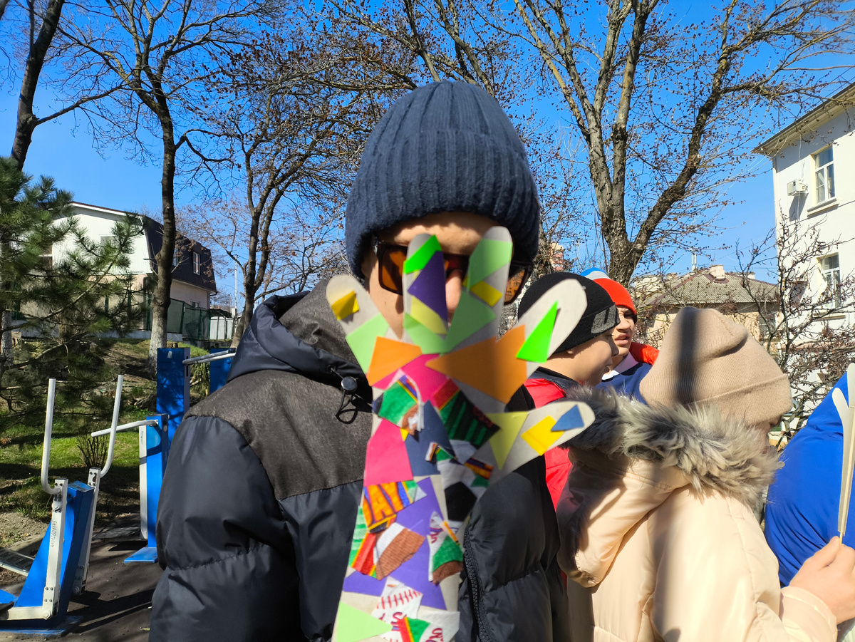 Тифлокомментарий. Гоша на улице с другими детьми. Лицо закрыто цветной ладошкой из картона