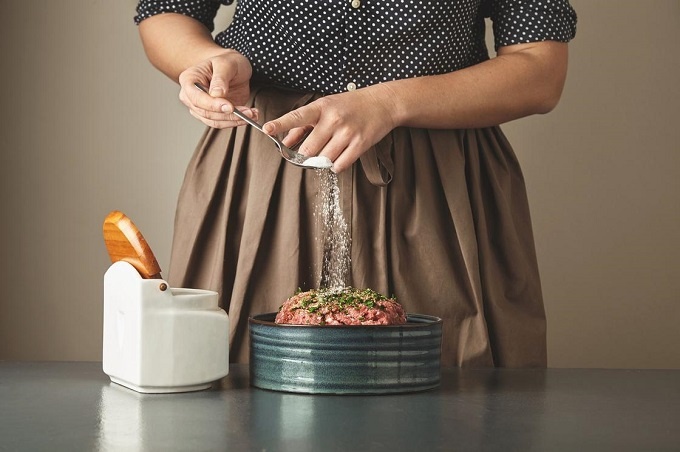 Housewife unrecognizable woman adds some salt in minced meat in ceramic bowl on blue table