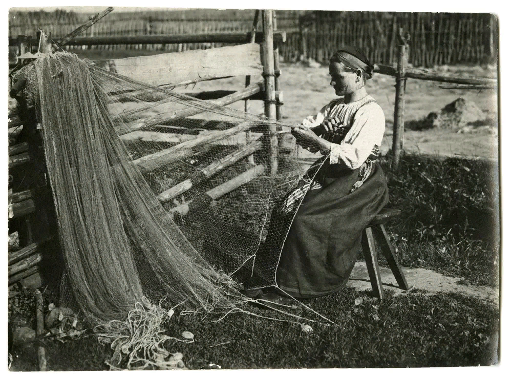 А на этом фото «Пожилая женщина сидит на самодельной скамеечке. На голове - повойник, белая рубаха, синий сарафан, подпоясанный синим пояском. В руке держит игличко, на среднем пальце - ножичек для обрезания нитки». Это тоже ижора, и сфотографирована она примерно там же, где и предыдущие рыбаки.