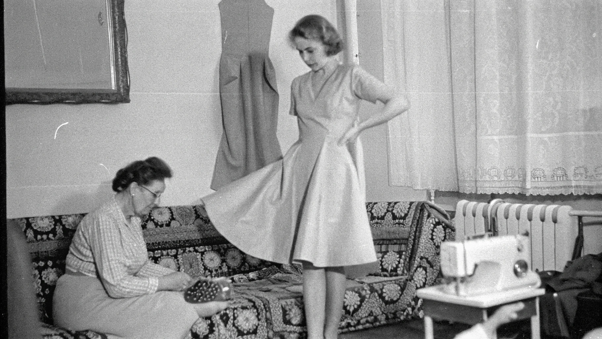 Примерка самодельного платья в домашних условиях. Молодая женщина стоит в элегантном платье с пышной юбкой, пожилая женщина на полу подшивает подол