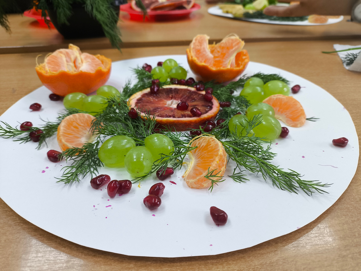 Тифлокомментарий. Белый картонный круг. Посередине кругляш апельсина, вокруг виноград, мандарины, зерна граната и укроп. 