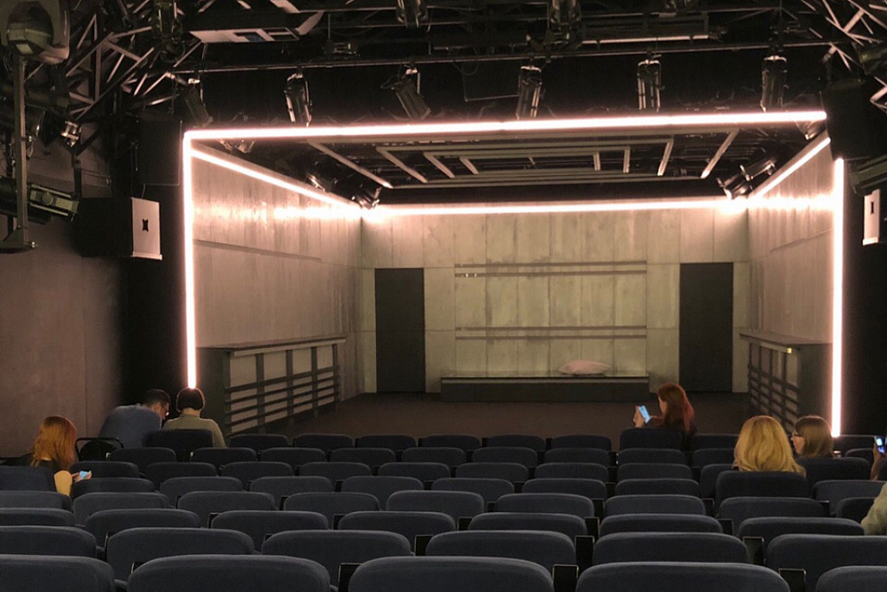 Сцена из спектакля «Преступление и наказание» (Приют комедианта, 2019). Богомолов убирает привычный бытовой антураж: стерильная серая коробка вместо петербургских интерьеров – визуальный символ отказа от «эмоциональной спекуляции» классического романа, оставляя лишь «экспозицию идей» героев​.