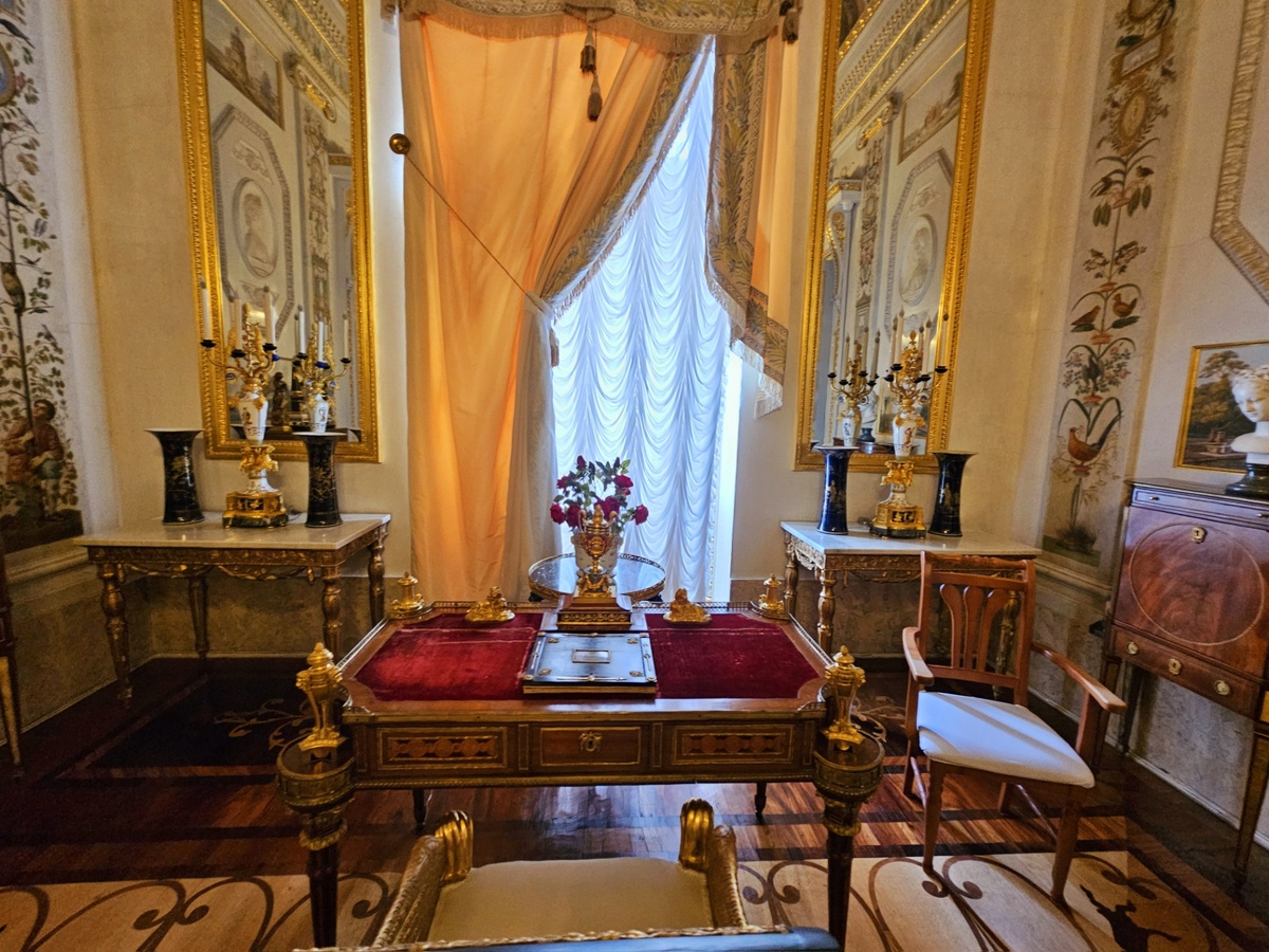 Против окна — письменный стол, периметр которого украшают часы-ваза с вращающимся циферблатом и четырьмя подсвечниками в виде ваз (Франция, мастер П. Денизо, 2-я пол. XVIII века). У стола — кресло с подлокотниками в виде крылатых сфинксов, выполнено специально для Будуара по проекту А. Н. Воронихина.