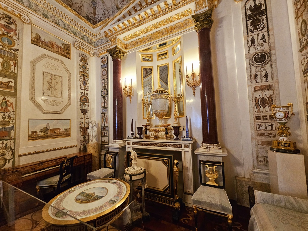 Между столом и камином – напольная ваза-курильница в виде треножника из разных сортов европейского мрамора (Италия, Б. Ферреро, 1780-е годы). Это подарок Павлу Петровичу от короля Сардинии Виктора-Амадея III. Слева от камина у стены стоит маленькое фортепьяно в корпусе наборного дерева. Оно было выполнено по заказу Екатерины II в Лондоне в 1774 году по рисункам английского архитектора Р. Адама. В углу между фортепьяно и камином находится мраморная группа «Амур и Психея», привезенная из путешествия (Италия, К. Альбачини, около 1780 года с античного оригинала). Справа от камина – фарфоровая ваза-часы, оправленная в бронзу (часовщик Л. Ж. Лагесс). К ней в ансамбль входят два канделябра (на противоположной стороне на консолях). Ансамбль был выполнен во Франции на Севрской фарфоровой мануфактуре в 1790-х годах, бронза мастерской П.-Ф. Томира.