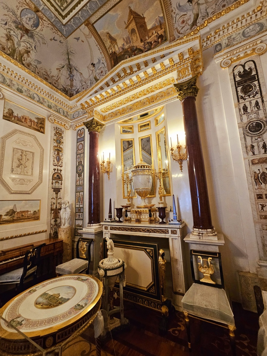 Между столом и камином – напольная ваза-курильница в виде треножника из разных сортов европейского мрамора (Италия, Б. Ферреро, 1780-е годы). Это подарок Павлу Петровичу от короля Сардинии Виктора-Амадея III. Слева от камина у стены стоит маленькое фортепьяно в корпусе наборного дерева. Оно было выполнено по заказу Екатерины II в Лондоне в 1774 году по рисункам английского архитектора Р. Адама. В углу между фортепьяно и камином находится мраморная группа «Амур и Психея», привезенная из путешествия (Италия, К. Альбачини, около 1780 года с античного оригинала). Справа от камина – фарфоровая ваза-часы, оправленная в бронзу (часовщик Л. Ж. Лагесс). К ней в ансамбль входят два канделябра (на противоположной стороне на консолях). Ансамбль был выполнен во Франции на Севрской фарфоровой мануфактуре в 1790-х годах, бронза мастерской П.-Ф. Томира.