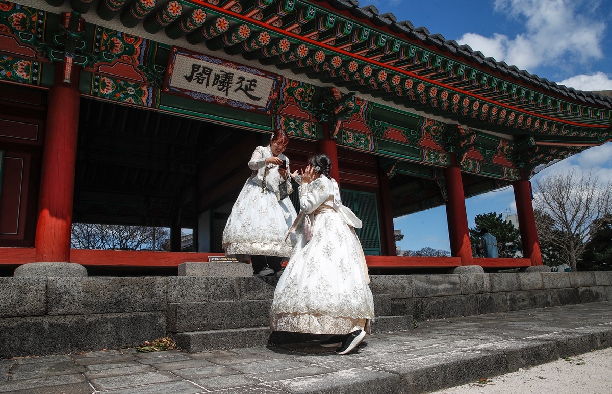 Иностранные туристы, одетые в корейскую традиционную одежду ханбок, фотографируются в Квандокчоне на острове Чеджудо 17 марта, месте съемок популярного сериала Netflix «Когда жизнь дарит тебе мандарины» (Newsis)