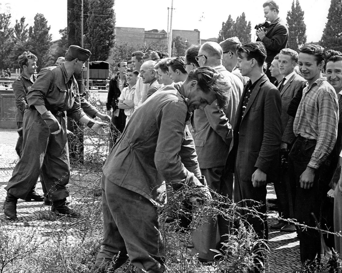 Восточногерманские солдаты, слева, устанавливают баррикады из колючей проволоки на границе, разделяющей Восточный и Западный Берлин, 13 августа 1961 года. Жители Западного Берлина, справа, наблюдают за работой.
