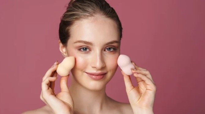 Молодая женщина с естественным макияжем демонстрирует два косметических спонжа разного цвета, прикладывая их к щекам, на розовом фоне