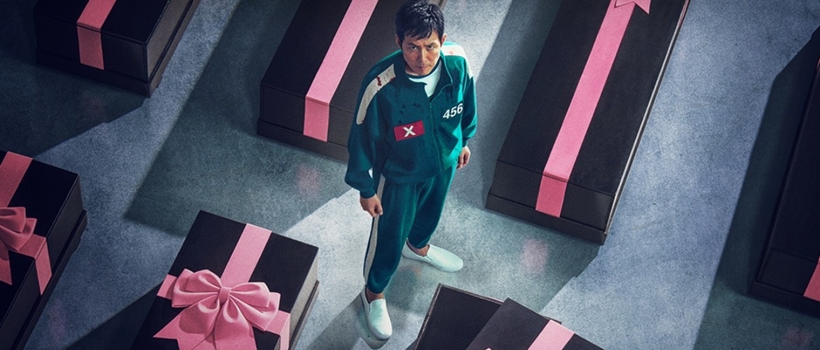 Сон Ки Хун (игрок 456) из Игры в кальмара 3 стоит в зеленом костюме среди черных ящиков, похожих на гробы, с розовыми бантами, вид сверху