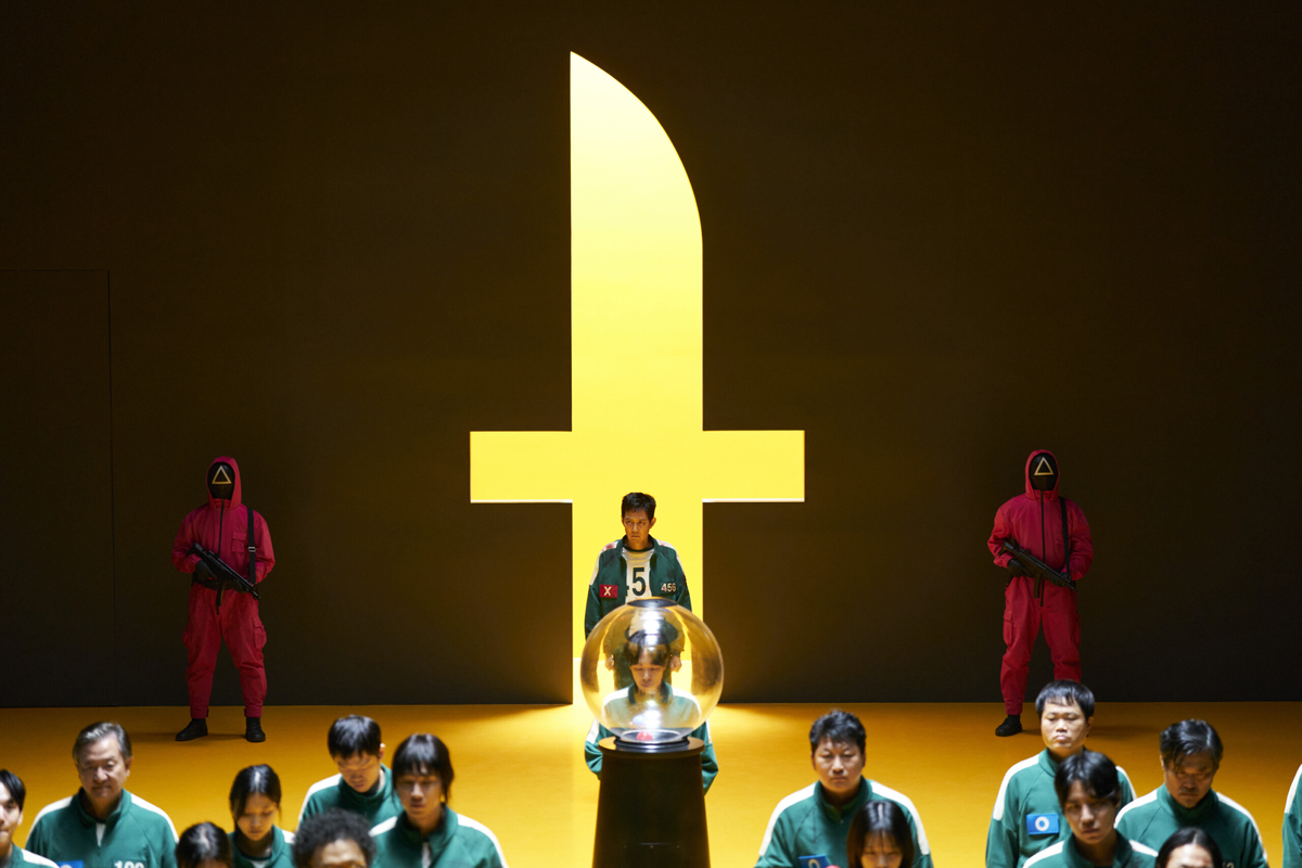 Игроки Игры в кальмара 3 в зеленых костюмах, Сон Ки Хун (456) в центре перед стеклянной сферой с золотой копилкой-свиньей, на фоне большой желтой светящейся фигуры и охранников в красном