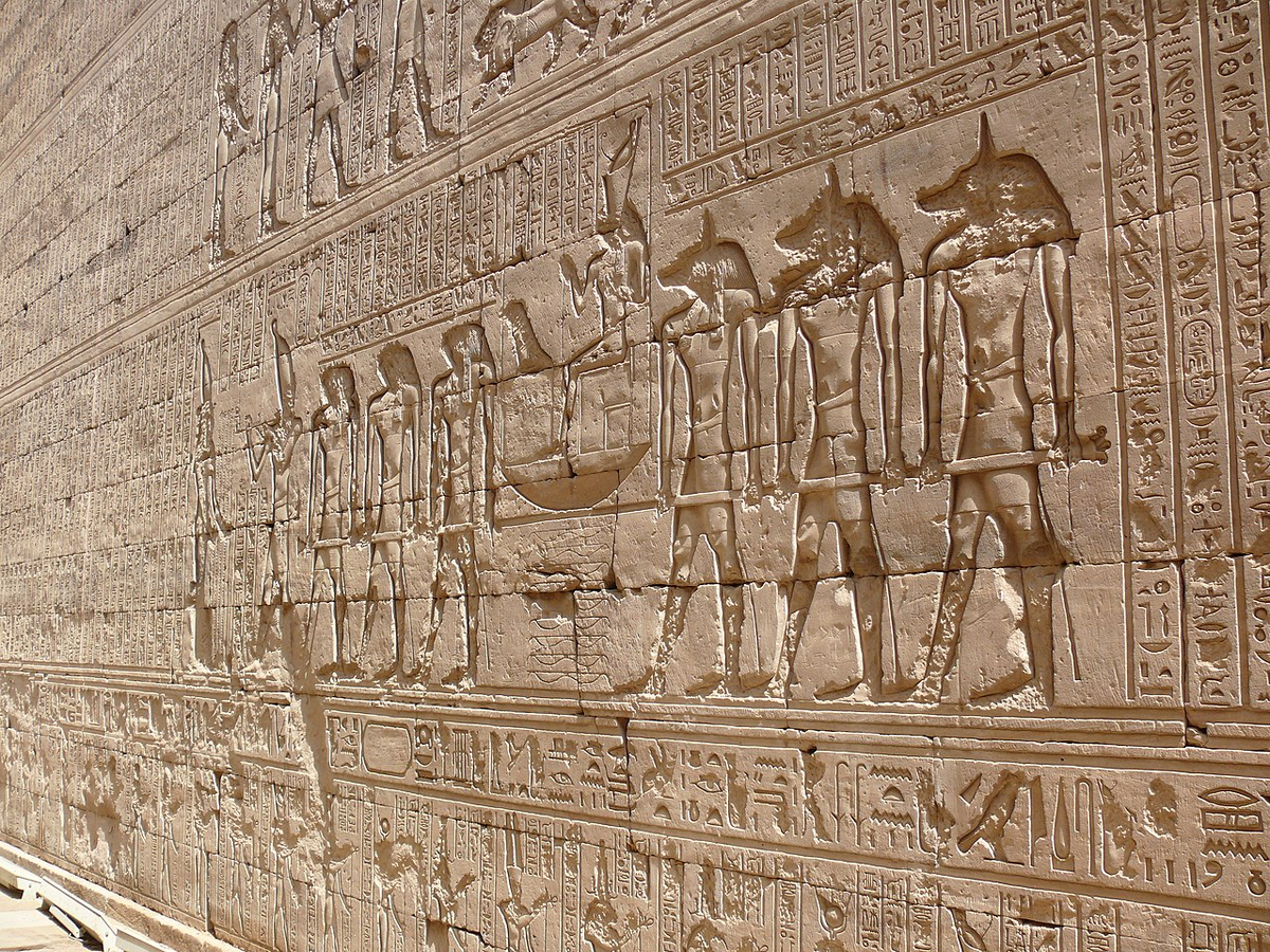 Рельеф из храма Эдфу в Египте: анубисоподобные фигуры сопровождают фараона, несущего двойную корону Верхнего и Нижнего Египта. Храм Эдфу посвящён богу Гору и считается одним из самых хорошо сохранившихся культовых комплексов античности. Этот сюжет часто интерпретируют как процесс инициации или «божественного воссоединения», когда правитель становился посредником между небом и землёй. Источник: Wikimedia Commons / автор Ad Meskens, свободная лицензия (Own work, CC BY-SA).