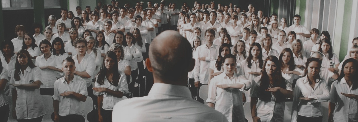 ГГ стоит спинной. Его ученики одетые в одинаковую одежду делают приветственный жест их движения. 