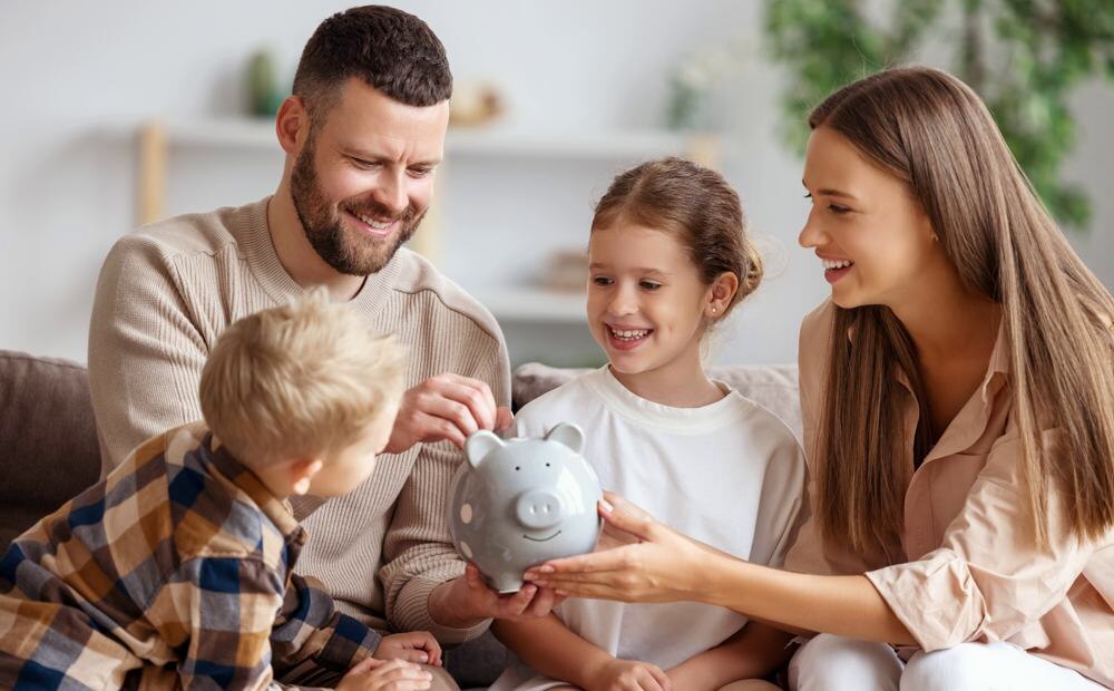      Happy family cheerful mother and father with kids smiling and putting coins into piggy bank while sitting on sofa at home tourist