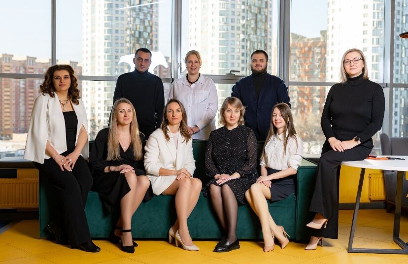 Тифлокомментарий. Рабочая команда Натальи. Мужчины и женщины одеты в деловом стиле кэжуал. Кто-то сидит на зеленом диване, а кто-то стоит за ним. На заднем плане — панорамные окна.