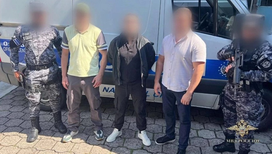    Мужчина, обвиняемый в убийстве петербуржца (в чёрном, по центру), в окружении сотрудников Интерпола и ФСИН перед экстрадицией, май 2025 года. Автор фото: Пресс-центр МВД России