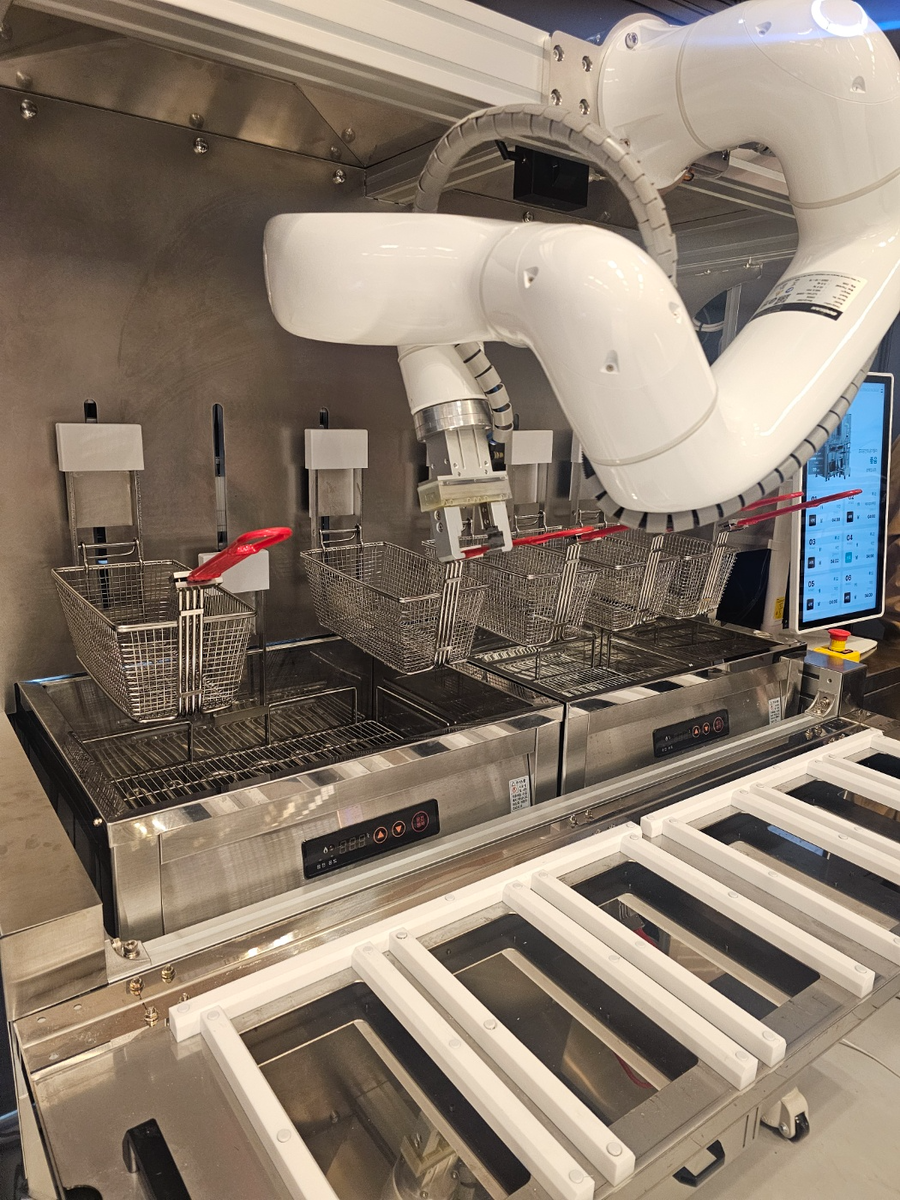 Робот для жарки, разработанный местным робототехническим стартапом itcobot, помещает контейнер во фритюрницу во время демонстрации на Ярмарке продовольственных услуг 2025 года в Coex на юге Сеула, 29 апреля (Чхве Чжэ Хи/The Korea Herald)