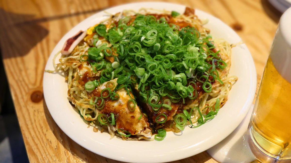 Тарелка хироимского окономияки, щедро украшенного зелёным луком, и холодный бокал пива на деревянном столе.