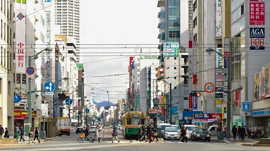 Современная улица в Хиросиме с трамваем посреди проезжей части, окружённая небоскрёбами, рекламой, машинами и прохожими.