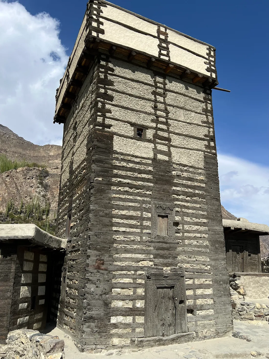 Деревянно-каменная сторожевая башня с резной дверью и окнами, а на заднем плане — горы.