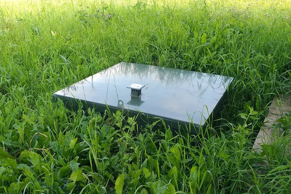 Герметичный пластиковый контейнер с трубами для сброса стоков и вывода очищенной жидкости. Закапывается в землю, а сверху — зеленая крышка, которая незаметна в траве