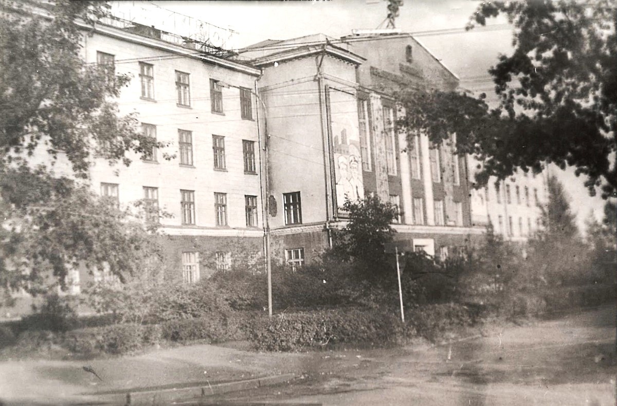 Фотография № 1. Так выглядел фасад главного корпуса КрасГМИ в годы нашей учебы - во второй половине 1980-х годов: невысокие кустарники и полуяблоньки, тополя, открытый проезд вдоль фасада здания.                                                                                                                                            
                                                                                                                                                                     
       В ризолите – выступающей части здания – на втором этаже располагались помещения деканатов трёх факультетов: лечебного, педиатрического, стоматологического. Крайнее дальнее окно ризолита на втором этаже – это окно кабинета нашего декана – декана лечебного факультета. До начала зимы 1984 года деканом лечфака был профессор, д.м.н. Лазаренко В.И., заведующий кафедрой глазных болезней. С весны 1985 деканом стал доцент, к.м.н. Турчина Т.К., доцент кафедры хирургических болезней № 2. На крыше и по центру здания видна двухметровая эмблема медицины – змея с чашей. Она изумрудно светилась вечерами всего несколько дней в году: на ноябрьские праздники, посвящённые Великой Октябрьской социалистической революции (а); на Новый год (б); на Праздник 1 Мая (в); на День Победы (г).
Кадр отснят от проезжей части улицы Партизана Железняка.
