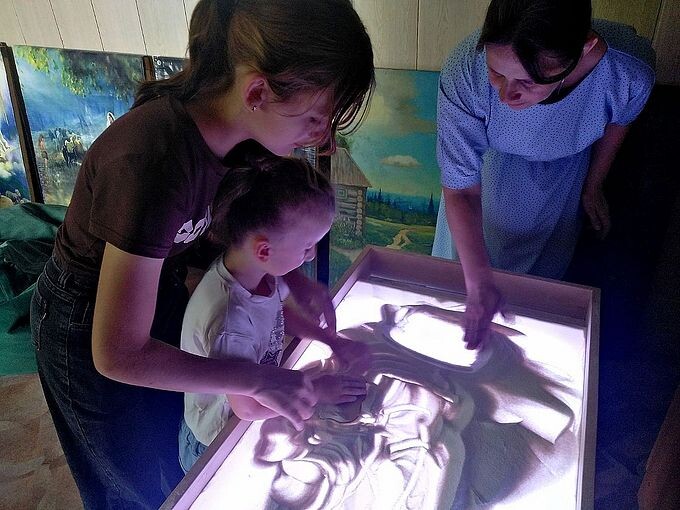 Мама и матушка Ирина показывают мальчику как рисовать руками на песке