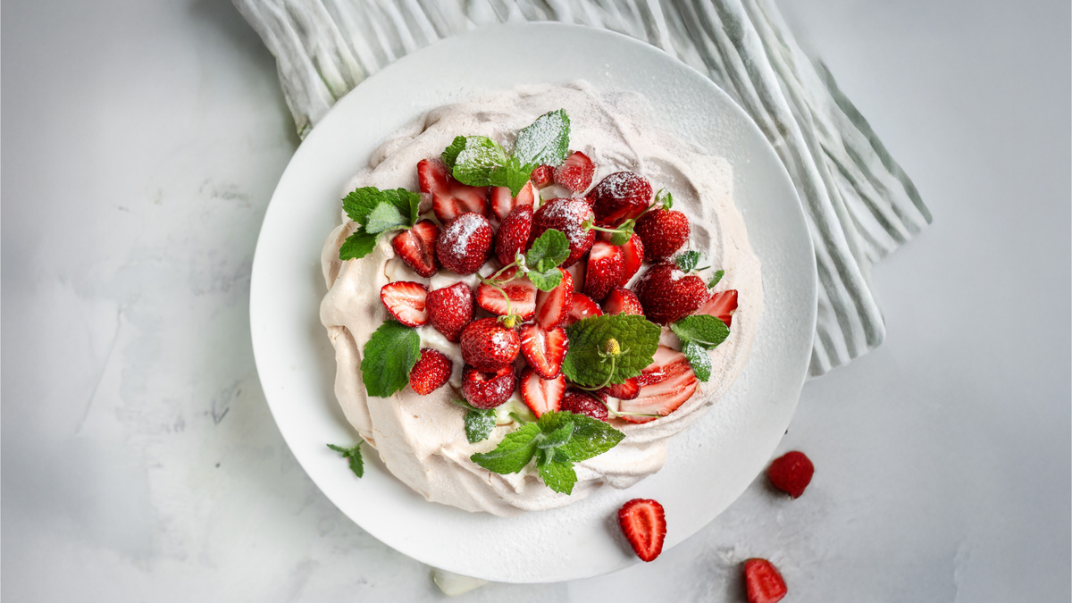 Готовый десерт «Павлова» с клубникой: хрустящая меренга со взбитыми сливками, украшенная свежими ягодами и мятой. Присыпана сахарной пудрой для элегантной подачи.