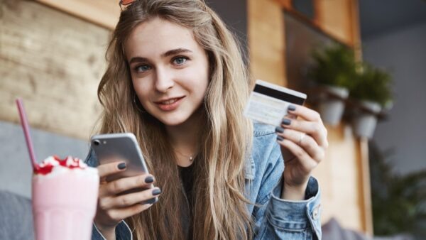    Eating out and candid people concept. Young blond woman paying for drink in cafe, using credit card and mobile phone, smiling at camera. Новостная служба "Правмир"