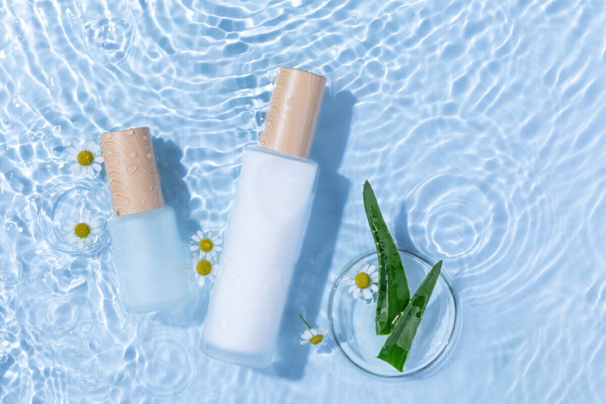    A top view of skincare bottles on a light blue water surface with aloe vera and daisy flowers Ксения Кузнецова