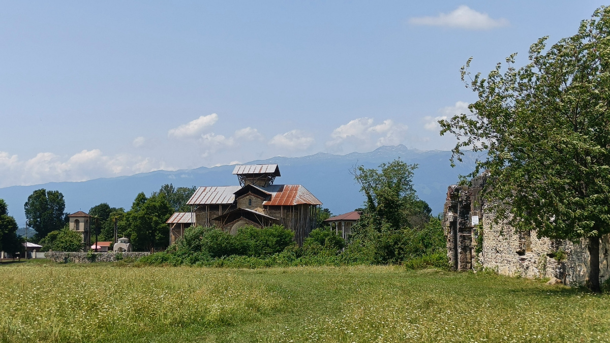 Лыхны. Вид на старинную Церковь Успения Пресвятой Богородицы (в центре кадра), часть стены руин дворца князей Чачба-Шервашидзе на фоне гор. Июнь 2025. Фото автора.
