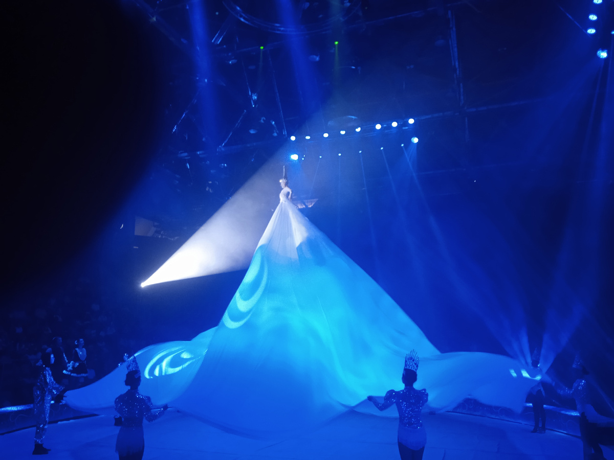 Вот такое огромное белоснежное платье на героине. Сама героиня висит под куполом цирка, а подол ее платья держат снизу со всех сторон. 