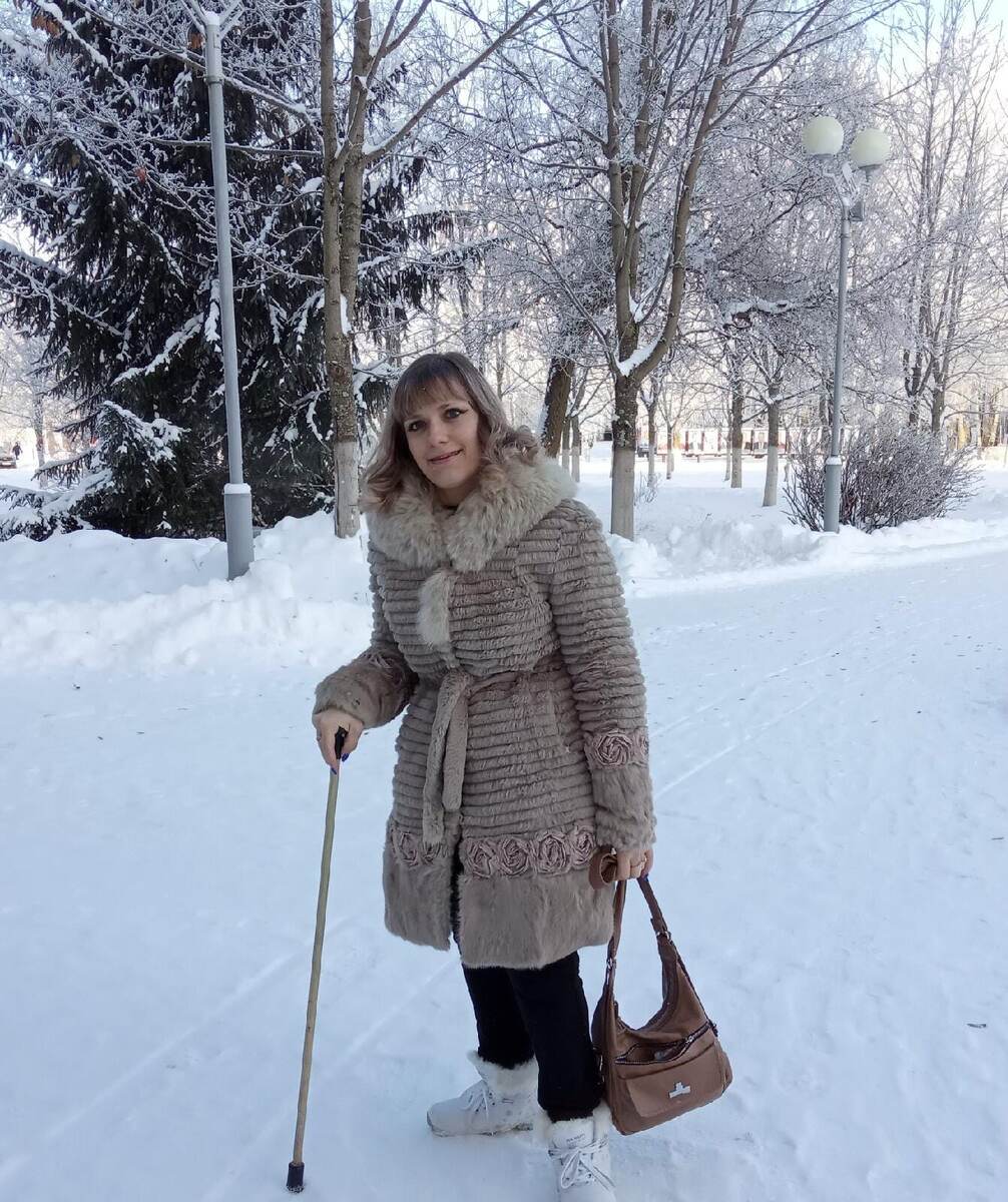 Тифлокомментарий. На фото улыбающаяся женщина в зимнем пальто с кружевными вставками, воротник оторочен серым мехом. Одной рукой она опирается на палочку, в другой руке — сумка. На заднем фоне — зимний лес, покрытый снегом.