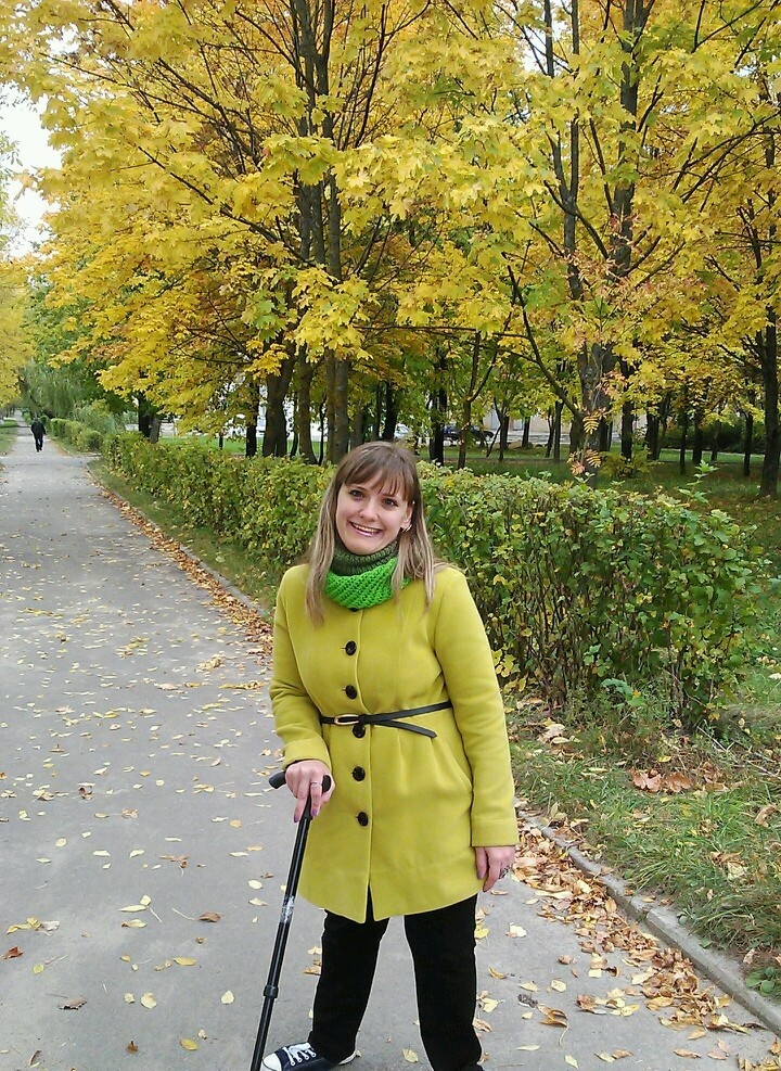Тифлокомментарий. На фото женщина в ярком салатном пальто опирается на палочку. На лице — широкая улыбка. На заднем фоне — кленовый лес с пожелтевшими листьями.