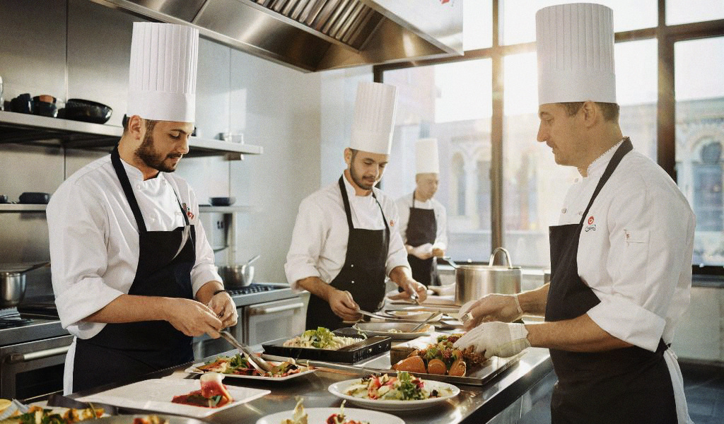 Повара в профессиональной кухне, которые готовят еду. Они одеты в белые поварские костюмы и черные фартуки. Один из поваров держит нож и нарезает ингредиенты на разделочной доске, другой повар накладывает еду на тарелку. В кухне видны различные кухонные принадлежности и оборудование.