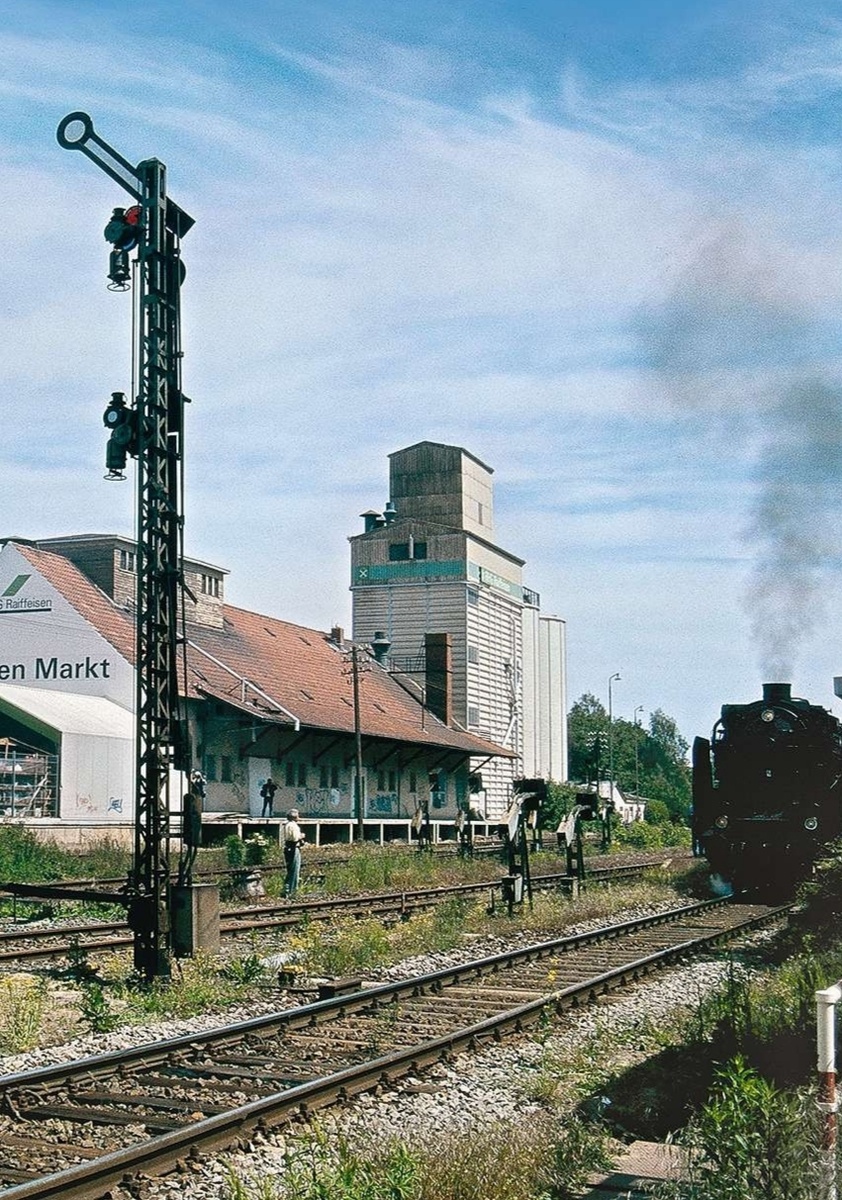 Raiffeisen Markt и современный зерновой склад с силосной башней на фоне, типичный вид для железнодорожных станций с сельхознагрузкой. На переднем плане — механический семафор и паровоз на путях