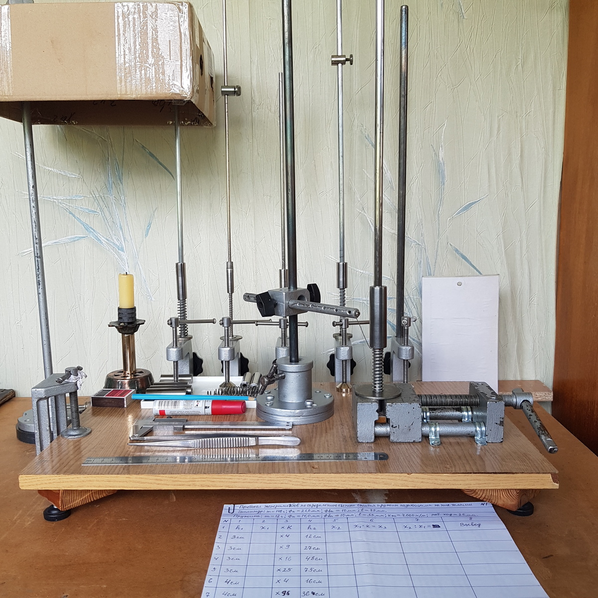 На переднем плане этого снимка вы видите подготовленный к заполнению черновик протокола проведения намеченной нами серии экспериментов. Далее вы видите само основание прибора с закрепленными на нем двумя основными вертикальными стойками. Одна из которых служит направляющей для падающего на пружину цилиндра. А другая служит для перемещения по ней держателя чертилки, которая отмечает максимальную степень сжатия пружины после падения на нее цилиндра. На заднем плане вы видите вспомогательные струбцины с закрепленными в них также основными вертикальными направлящими, только меньшего диаметра. С соответсвующими им пружинами и падающими на них грузами. Также на поверхности этого прибора вы видите свечу, в пламени которой боковые поверхности цилиндров покрываются копотью. И только часть приборов и измерительных инструментов, которые применяются при проведении данных экспериментов.