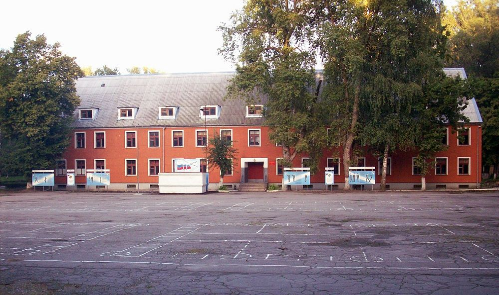 Фотоиллюстрация: 1971. Город Пионерский Калининградской области. 9-й Флотский экипаж ДКБФ (в/ч 87284 "Б"). Тот самый строевой плац, на котором наше отделение "вышаков", Сергей из Тбилиси и наш двухметровый Ваня Пецко отрабатывали приёмы строевого шага, поворотов и построений. На плацу размечены белой краской места расположения матросов в строю, места поворотов, коробки для расположения отделений и т.д. На стендах плакаты с описанием строевых приёмов. С этой трибуны командир полка, гвардии капитан 1 ранга принимал строевые смотры. Здание позади - это главное административное здание 9-го Флотского экипажа, место размещения кадровых военнослужащих полка, канцелярия, штаб и т.д. Фото сделано летом, но всё было точно также и в ноябре-декабре 1971 года.