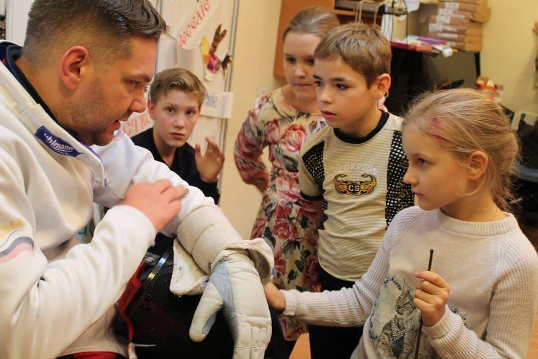 Тифлокомментарий. Николай рассказывает что-то детям, показывая на предплечье. Дети держат его за руку и с интересом смотрят на него. На Николае белая форма и перчатка на левой руке.