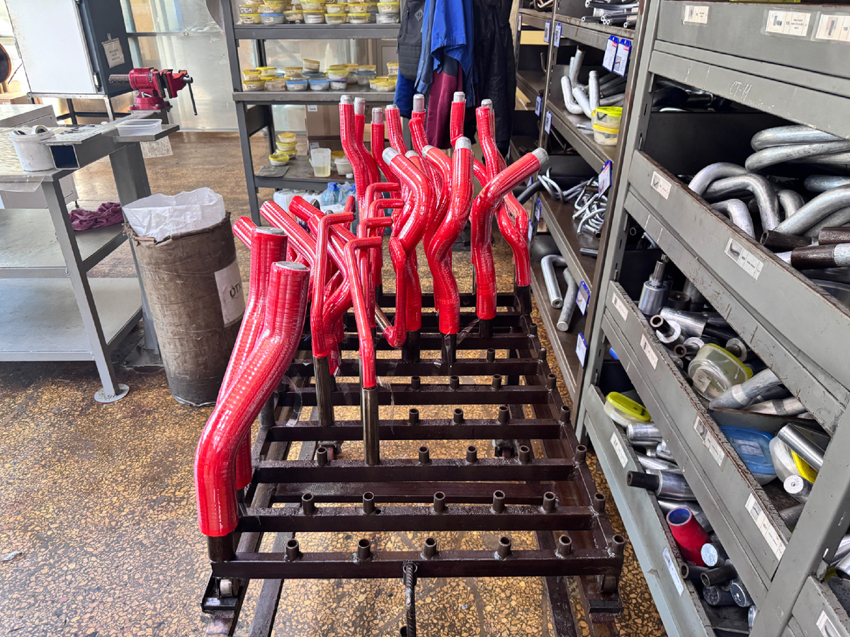 партия готовых силиконовых патрубков сложной формы от ПТП, установленных на сушильной стойке после формовки. Изделия армированы кордовой нитью, имеют пятислойную структуру и характерную глянцевую защитную плёнку. Эти патрубки рассчитаны на установку в ограниченных подкапотных пространствах (например, LADA GRANTA, KALINA, VESTA), где требуются точные геометрические изгибы. ПТП использует прочные металлические шаблоны (видны на заднем плане) для сохранения формы в процессе вулканизации — это гарантирует отсутствие заломов и стабильную проходимость канала даже при высоких температурах до +270 °C и давлении до 5 атмосфер.