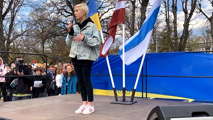    Хаматова выступила на "антивоенном митинге" в Риге на фоне трёх флагов - украинского, латвийского и... бело-сине-белого флага российской оппозиции. Скриншот: видео в vk.com