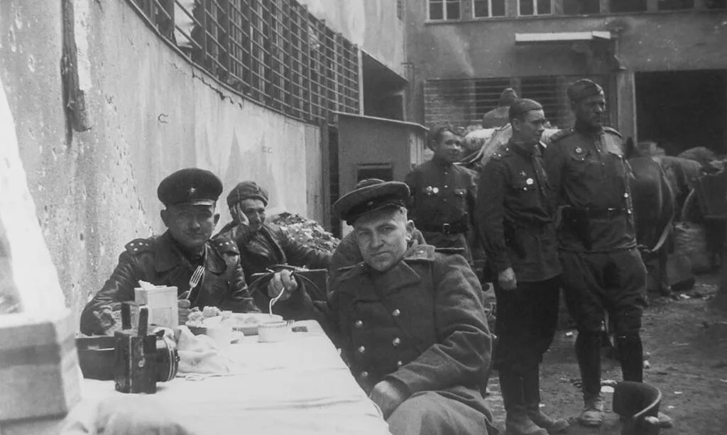 Историческое фото советских солдат, отдыхающих перед штурмом Берлина в 1945 году. На изображении они сидят за столом с полевыми принадлежностями – чашками, котелками, снаряжением; видно выражение усталости и сосредоточенности, как перед решающим моментом