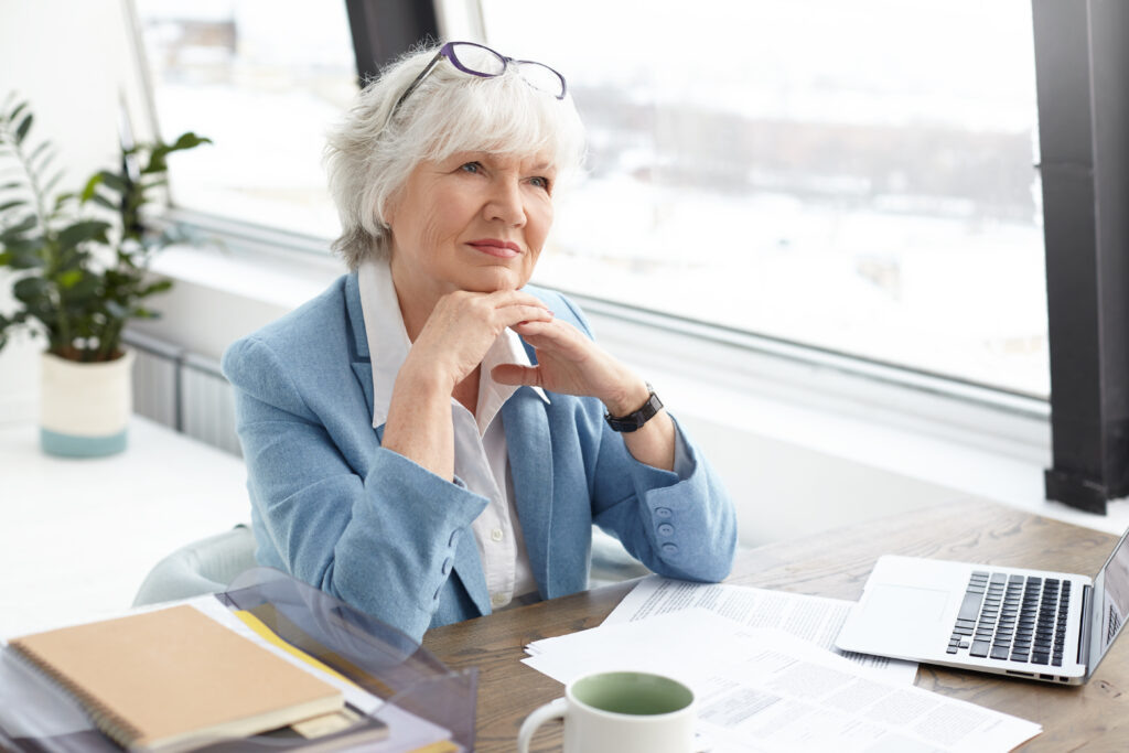    Succesfful skilled attractive elderly female editor of popular fashion magazine sitting at her workplace with papers, mug and open portable computer, clasping hands, having pensive facial expression Журналист