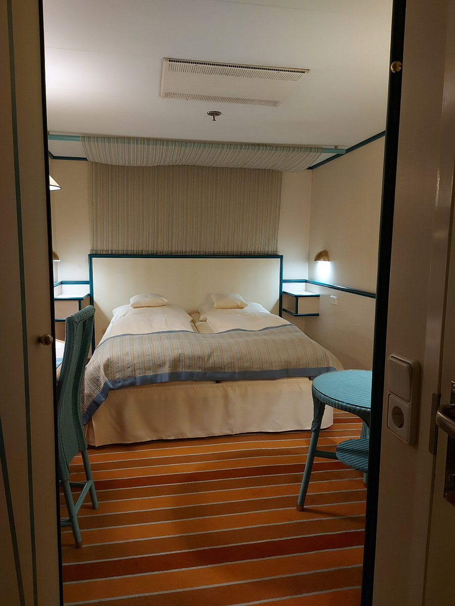 Каюта при заезде: свет приглушён, ковролин полосат, всё аккуратно. На кровати - не я, пока что 😄Кстати, кондиционер всегда работает, в ноль выключить нельзя, только регулировать, больше-меньше.