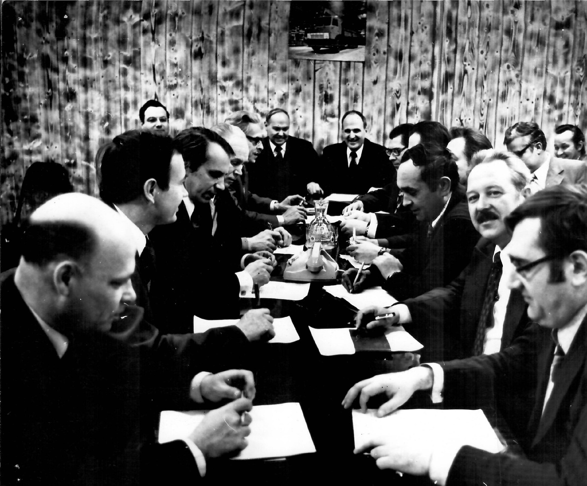 За столом сидят слева направо: 1. Захваткин - главный сварщи КАМАЗа. 2. Кузнецов Юрий Гаврилович – директор ремонтно-инструментального завода. 3. …  4. Тананин Александр Николаевич - главный металлург КАМАЗа. 5. Клепацкий Борис Трофимович – главный инженер КАМАЗа. 6. Ивженко Владимир Яковлевич – директор автомобильного завода. 7. Плахов Анатолий Максимович – технический директор КАМАЗа. 8. Шуваев Александр Иванович - заместитель главного инженера КАМАЗа по подготовке производства. 9. … 10. … 11. Гужавин - начальник ОТК автомобильного завода. 12. Колышкин - заместитель главного металлурга КАМАЗа.. 13. Остроушко Юрий Андреевич - начальник экспериментального цеха УГК КАМАЗа.