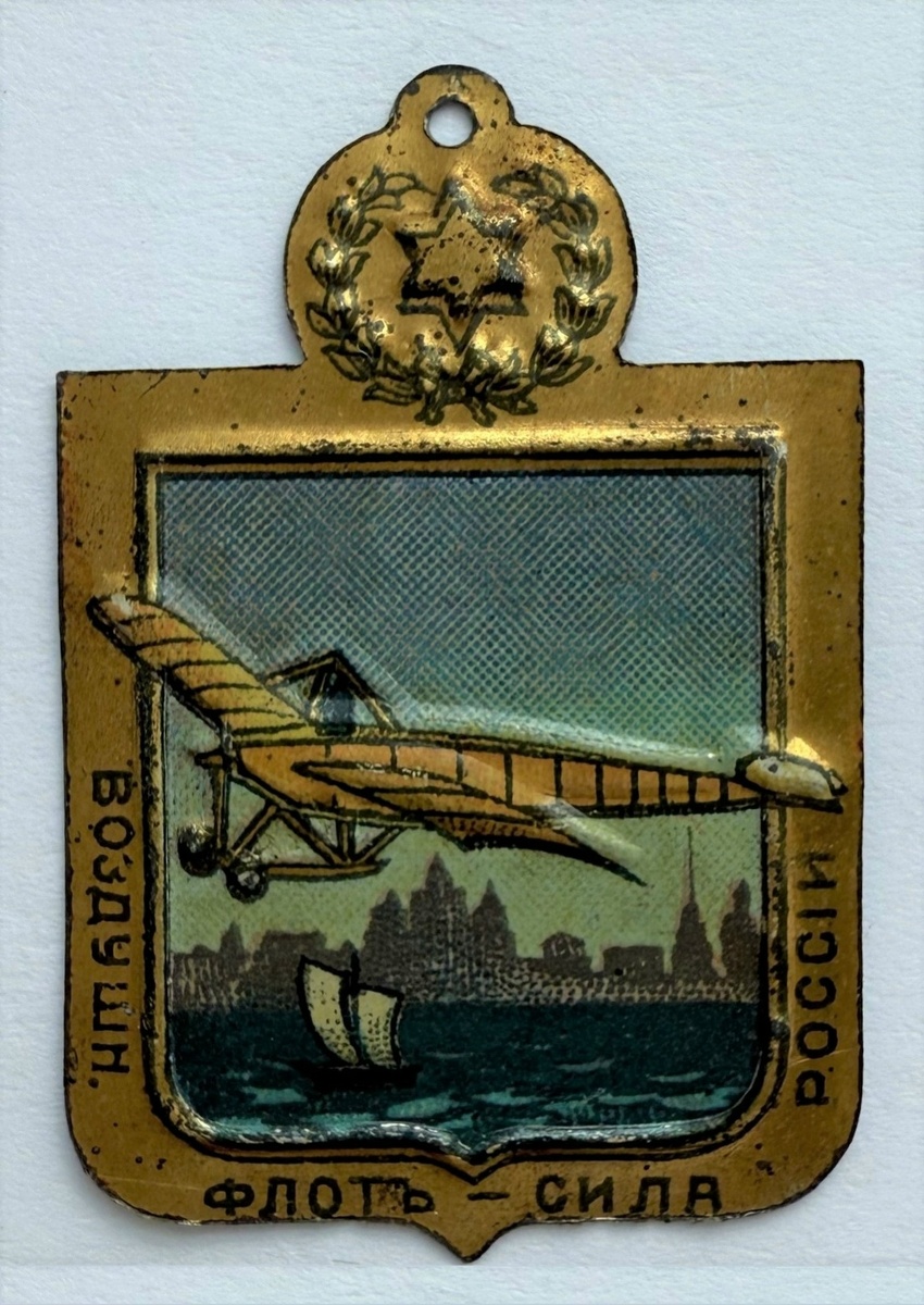 ОГС КП-336
Жетон выполнен в форме фигурного щита. В верхней части - круг с ушком, внутри которого расположена шестиконечная звезда и лавровый венок. По краю - жёлтый ободок с надписью: «ВОЗДУШН. ФЛОТЪ – СИЛА РОССIИ». В центре на фоне голубого неба - аэроплан, под ним - акватория, лодка с парусами, на дальнем плане - силуэты городских построек. Название фабрики-изготовителя на оборотной стороне: «Т-во Жесть РЕВЕЛЬ».