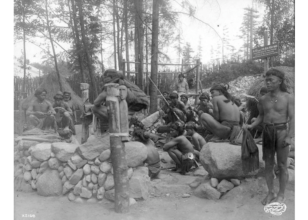 Организаторы Аляско-Юконско-Тихоокеанской выставки стремились создать на прогулочной улице Pay Streak «аутентичную» и одновременно развлекательную экспозицию — деревню игоротов. На этом фото изображена группа мужчин-игоротов перед каменным строением с табличкой «A-TO» и «PA-BA-FOO-NAN», обозначающей его как место собраний для мужчин.