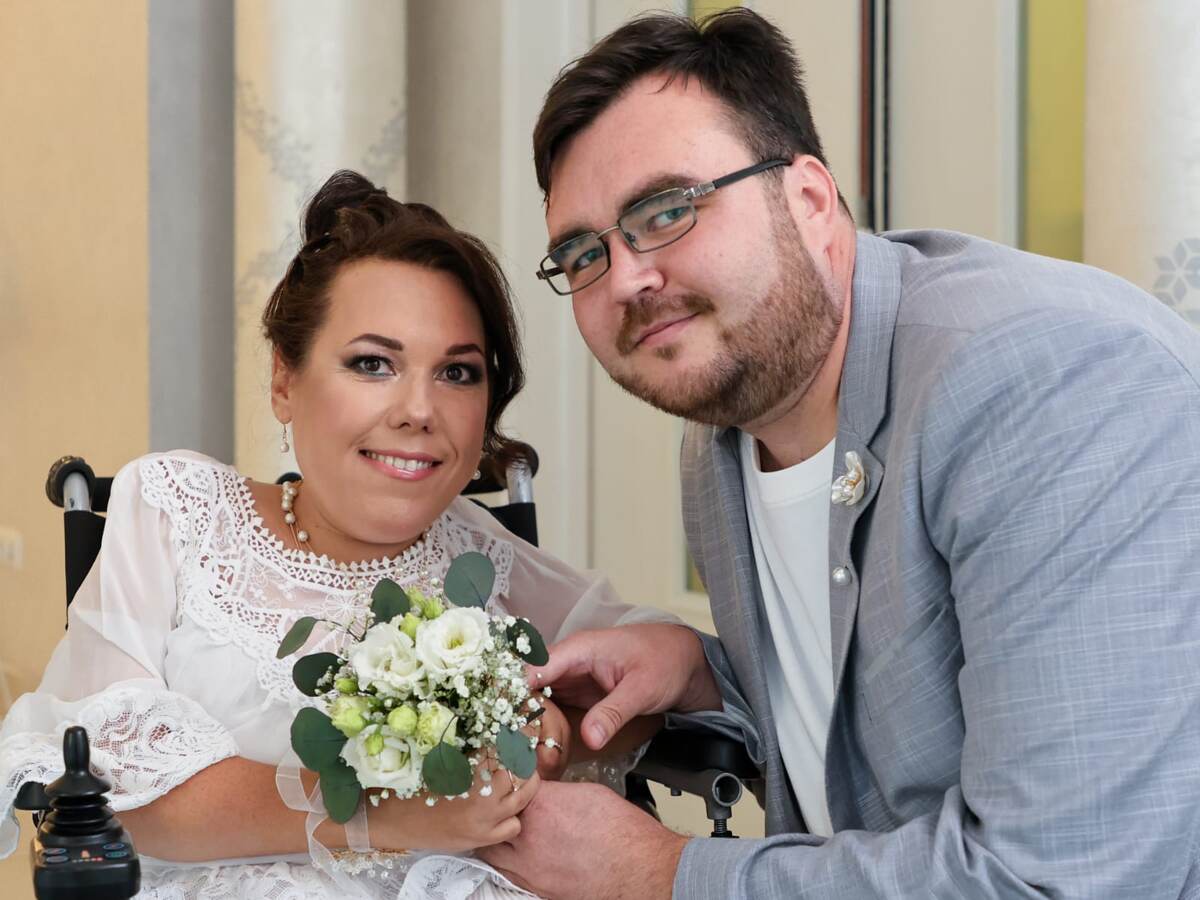 Тифлокомментарий. Татьяна с мужем на регистрации брака. Татьяна в белом кружевном свадебном платье с букетом белых роз в руках. Женщина в коляске. Муж стоит рядом, на нем серый пиджак с бутоньеркой и белая футболка. Фото: личный архив Татьяны Горбаневой.